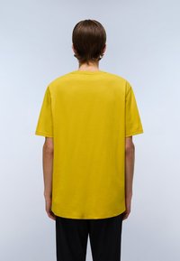 Person med kort brunt hår, iført en ensfarvet gul t-shirt med korte ærmer og sorte bukser, står med ryggen til i et lyst miljø.