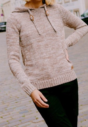 Person standing outdoors wearing a beige knitted hoodie with drawstrings and black pants, hand resting on hip and partially visible blond hair.
