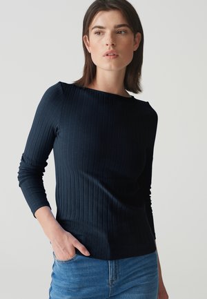 Long-sleeved ribbed top in dark navy fabric, featuring a boat neckline and a fitted silhouette. Paired with light blue denim jeans.