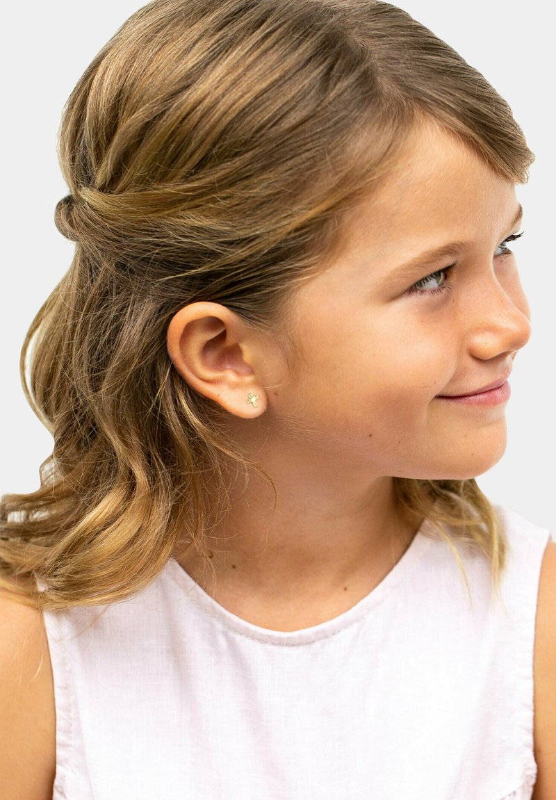 Boucles d'oreilles en fleurs dorées sur une fille aux cheveux ondulés mi-longs, portant un haut rose clair. Subtil éclat des boucles d'oreilles.