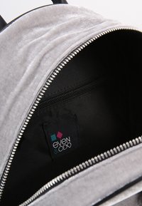 Interior de una mochila de terciopelo gris con un bolsillo con cremallera y un forro negro. Presenta una etiqueta de logo con un diseño geométrico.