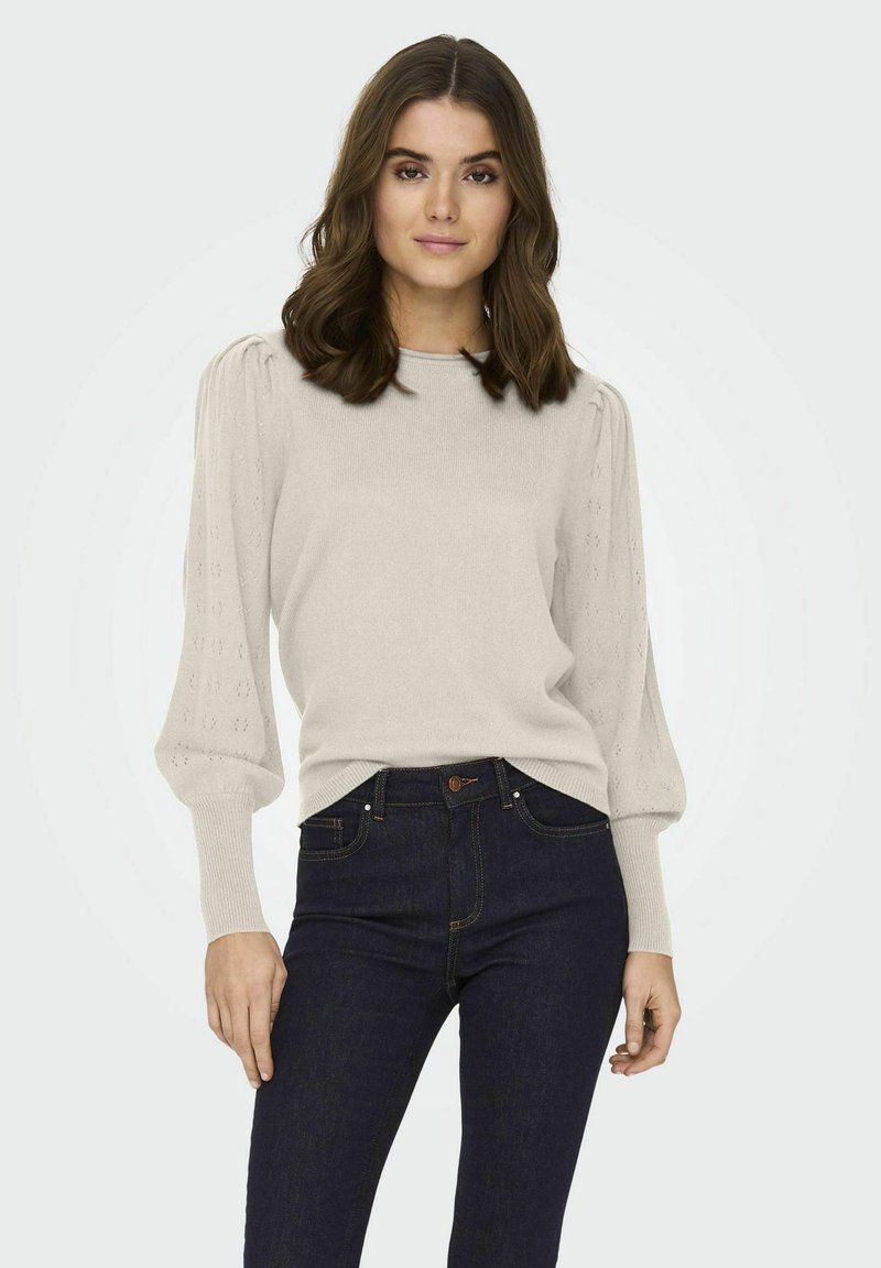 Suéter de punto beige claro con mangas abullonadas texturizadas, puños acanalados y escote redondo. Combinado con jeans azul oscuro.