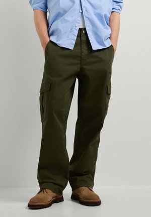 Persona vistiendo pantalones cargo verde oliva, zapatos marrones y una camisa azul claro con las manos en los bolsillos, de pie frente a un fondo simple.