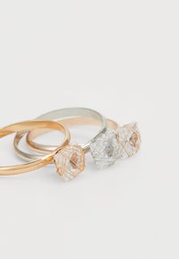 Tre anelli: due bande metalliche semplici in oro e argento, più uno con una pietra trasparente avvolta in filo di oro rosa, con forme geometriche.
