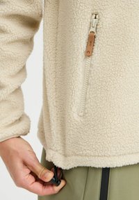 Pull-over en polaire beige avec une surface texturée, une poche zippée avec tirette en cuir, et un cordon de serrage réglable à l'ourlet.