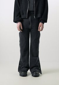 Sorte denimbukser med en frontlynlås, brede flagrende ben og trykknapdetaljer på siden. Båret med en matchende sort cropped jakke og kraftige sorte støvler.