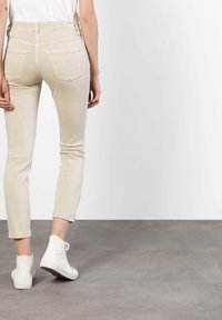 Beige avkuttede jeans med en tettsittende design, som har baklommer og frynsete kanter, kombinert med hvite høye sneakers.