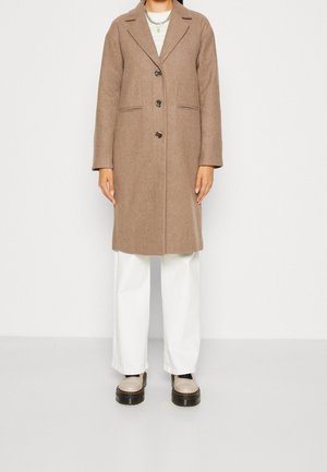 Personne portant un manteau brun boutonné jusqu'aux genoux par-dessus un pantalon blanc à jambes larges et des chaussures compensées beiges, se tenant devant un fond blanc uni.