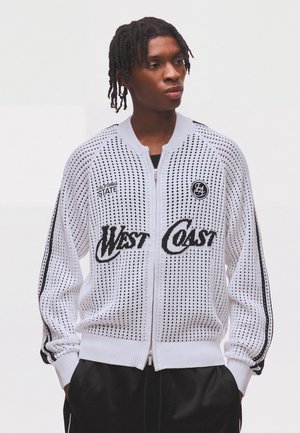Eine junge Person mit geflochtenem Haar trägt eine weiße Netzjacke mit Reißverschluss mit dem Text "West Coast" und schwarzen Streifen an den Ärmeln.