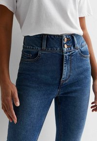 Jeans de denim azul de talle alto con cierre de dos botones, diseño de cinco bolsillos y detalles de costura a lo largo de las costuras. Textura suave.