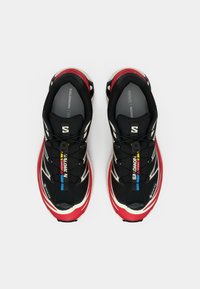 Schwarze und rote Sportschuhe mit einem Obermaterial aus Mesh, skulpturalem Design, strukturierten Sohlen und kontrastierenden gelben und blauen Akzenten.