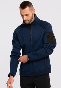 Niebieska kurtka zapinana na zamek z fakturowanej tkaniny, wysoki kołnierz, czarna kieszeń na rękawie oraz dwie kieszenie boczne zapinane na zamek. Kontrastowe przeszycia przy mankietach.