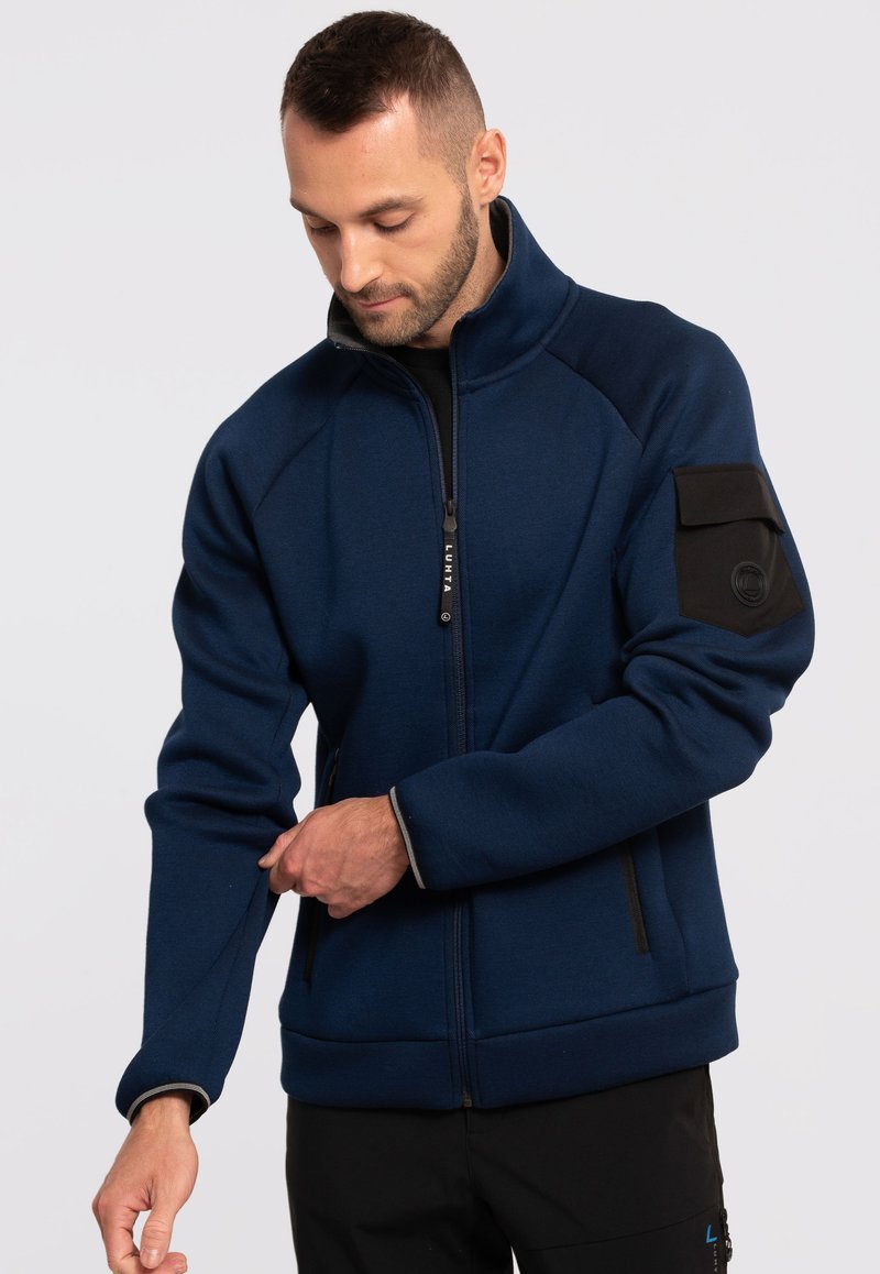 Niebieska kurtka zapinana na zamek z fakturowanej tkaniny, wysoki kołnierz, czarna kieszeń na rękawie oraz dwie kieszenie boczne zapinane na zamek. Kontrastowe przeszycia przy mankietach.