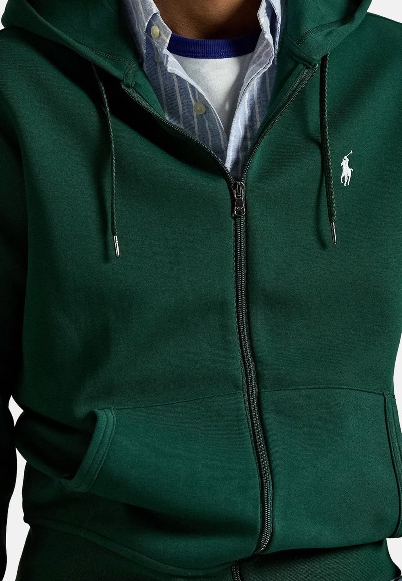 Sudadera verde con cremallera y capucha ajustable con cordón, bolsillos frontales y un pequeño logo blanco. Está hecha de una tela suave con un diseño ajustado.