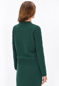 Pulover verde tricotat cu mâneci texturate, din tricot cu cablu, accentuate de mărgele mici. Talie ajustată și manșete ribbate.