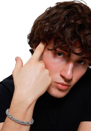 Bracelet en chaîne argentée avec des maillons complexes, porté au poignet d'une personne aux cheveux bouclés, la main positionnée près du visage, sur un fond uni.