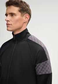 Zwarte sportjack met hoge kraag, met grijze patroonaccenten op de schouders. Gladde textuur, rits aan de voorkant en een aansluitend ontwerp.