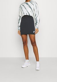 Zkrácená mikina s batikováním v bílé a zelené barvě, spárovaná s černou mini sukní s jemným bílým logem, přes kterou jsou obuty bílé tenisky.