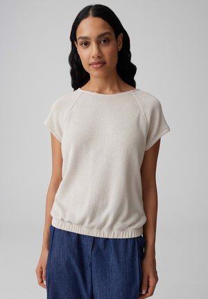 Frau mit langen dunklen Haaren, die ein beige kurzärmeliges Oberteil und blaue Hose trägt, steht vor einem einfachen hellgrauen Hintergrund.