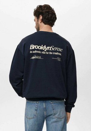 Man with dark curly hair wearing navy sweatshirt with "Brooklyn Sense" text and city skyline graphic, paired with light blue jeans, facing away.