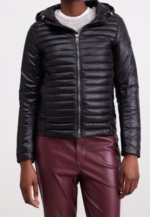 Person iført en sort quiltet jakke med lynlås og hætte over en lysegrå sweater og vinrøde læderbukser.