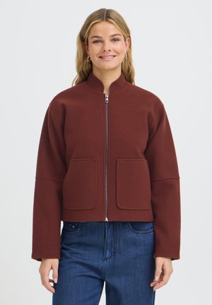 Femme souriante, portant une veste bordeaux à fermeture éclair avec de grandes poches à l'avant et un jean bleu, debout devant un fond blanc uni.