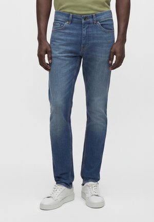 Άνδρας που φοράει μπλε τζιν παντελόνι slim-fit και λευκά αθλητικά, στέκεται μπροστά σε απλό φόντο.