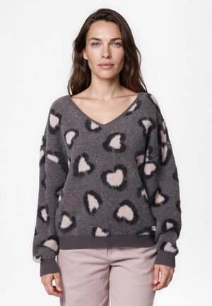 Femme aux longs cheveux bruns portant un pull gris à col en V avec des motifs de cœurs roses et noirs et un pantalon rose clair, debout devant un fond uni.