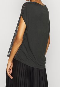 Černý top s krátkým rukávem a kulatým výstřihem, s patterned bočním panelem zdobeným tečkami v žluté, modré a šedé barvě na tmavém pozadí.