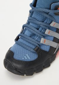 Blue textured mesh upper with black rubber sole, gray straps, and orange accents. Features three stripes logo near the toe.