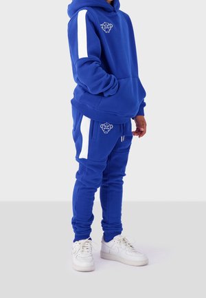 Royalblaue Kapuzenjacke und Jogginghose-Set mit weißen Seitenstreifen und Logo. Mit Kängurutasche und gerippten Bündchen. Kombiniert mit weißen Sneakers.