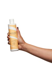 Fles curl conditioner vastgehouden in hand. De fles is cilindrisch, wit met een textuur gele label met een golvend ontwerp en zwarte tekst.