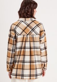 Camisa de cuadros de manga larga en tonos marrón, blanco y negro. Presenta un canesú con pliegues, puños con botones y una tela suave y texturada.