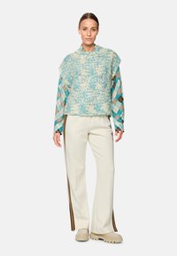 Turquoise knitted sweater with a textured surface, layered over a plaid patterned shirt. Cream wide-leg pants feature side stripes and chunky shoes.