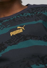 T-shirt Puma con strisce astratte in navy, teal e nero, caratterizzato da un logo dorato sul petto. Tessuto morbido con scollo a girocollo.