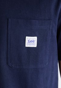 T-shirt en coton bleu marine avec une poche poitrine carrée comportant une étiquette blanche avec un texte violet. Le tissu a une texture lisse et un design côtelé.