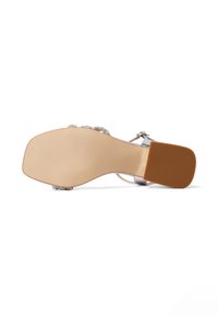 Sandal aperto con un design di cinturini argentati, decorata con decorazioni; presenta una suola liscia color tan e un tacco leggermente testurizzato.