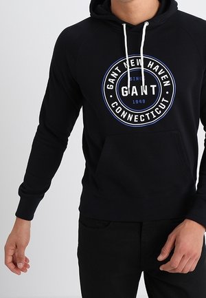 Mężczyzna noszący czarną bluzę z kapturem z białymi sznurkami i okrągłym logo GANT New Haven Connecticut na piersi.