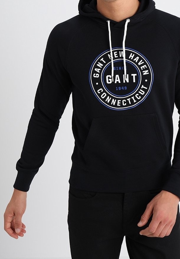 Homme portant un sweat à capuche noir avec des cordons blancs et un logo circulaire GANT New Haven Connecticut sur la poitrine.