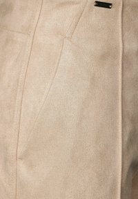 Beige mockaklänning med sömdetaljer och en liten svart metallmärke fäst nära övre kanten.