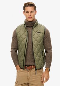 Gequilted olijfgroen vest met een hoge kraag, ritsluiting en zijzakken, gedragen over een bruine coltrui. Beige broek maakt de look compleet.