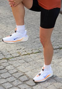 Białe buty sportowe z pomarańczowymi akcentami i niebieskim logo, z teksturowaną podeszwą i dopasowaniem przypominającym skarpetę, noszone z czarnymi szortami i białymi skarpetkami.