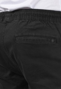 Black pants made of smooth fabric, featuring an elastic waistband and a small, flat back pocket with a button detail.