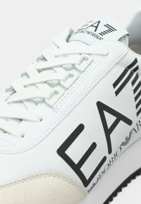 Fehér sportcipő fűzős záródással, sima textúrával, hangsúlyos fekete márkajelzéssel és krémszínű szarvasbőr díszítéssel.