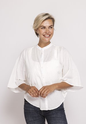 Blonde Frau lächelnd und nach rechts schauend, trägt eine lockere weiße Bluse mit weiten Ärmeln und dunkle blaue Jeans, steht vor einfachem Hintergrund.