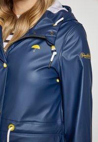 Tamsiai mėlyna vandeniui atspari striukė su blizgiu paviršiumi, geltonomis detalėmis, reguliuojama raištelių kapisonu ir mažas skėčių logotipas ant krūtinės.