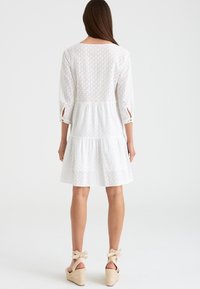 Vestido blanco de broderie anglaise con mangas de tres cuartos, diseño en capas y aberturas en los extremos de las mangas. Combinado con alpargatas beiges.