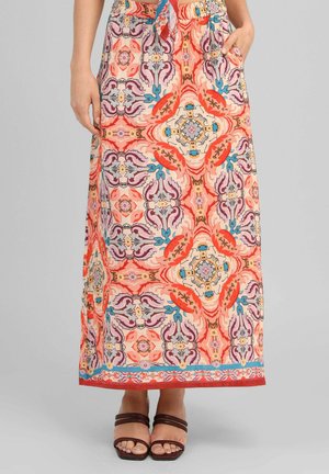 Femme portant une jupe longue colorée et à motifs avec des poches latérales et des sandales à brides marron sur un fond uni.