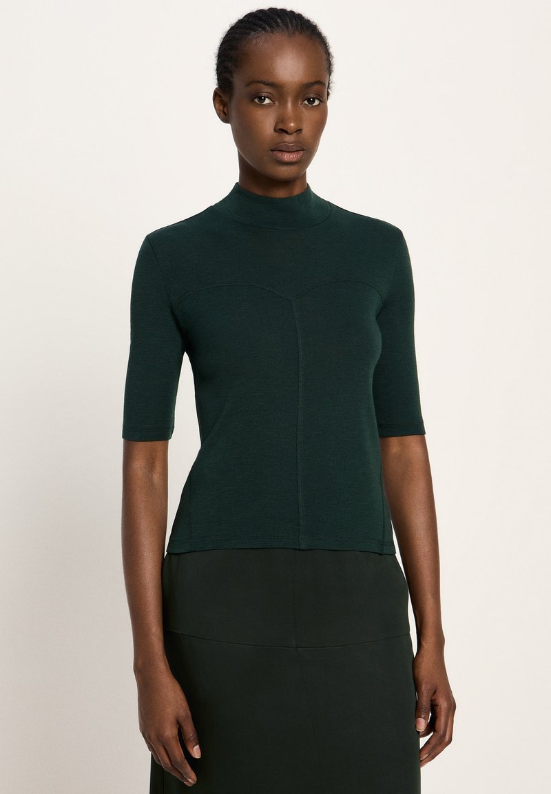 Woman with braided hair wearing a dark green short-sleeve high-neck top and matching dark skirt against a plain beige background.