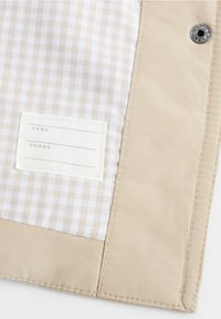 Beżowa saszetka o teksturowanym wykończeniu, z jasną kraciastą podszewką i materiałową metką na imię i telefon, zabezpieczona metalowym zatrzaskiem.